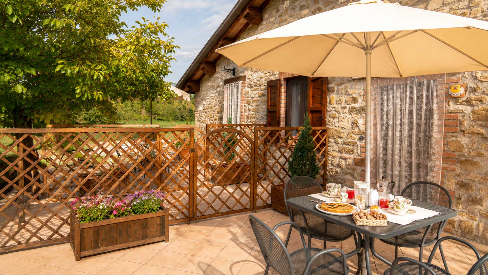 Terrazza del''appartamento Girasole ina agriturismo umbro Domus Santa Croce.  Colazione all'aperto con ombrelloni, tavolo e sedie in ferro, immersa nel verde e fiori.